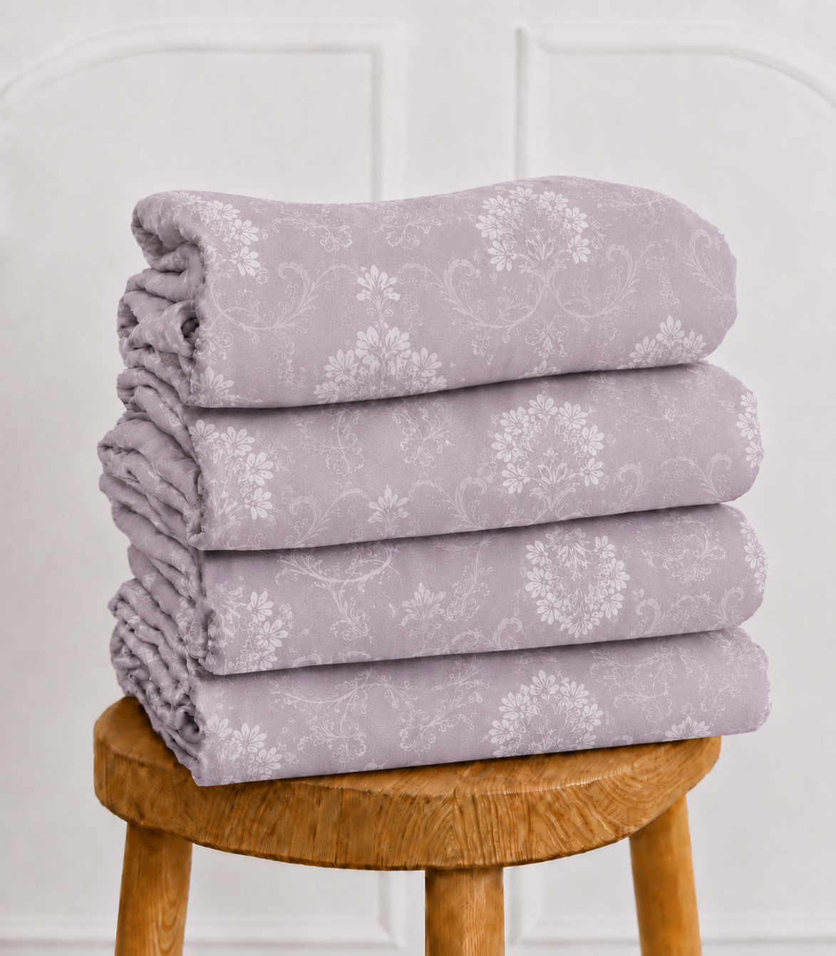 Folded king size misty lilac cotton bedsheet with delicate white floral vine pattern neatly stacked on a wooden stool. Premium fabric offers softness, durability, and a sophisticated botanical design.
