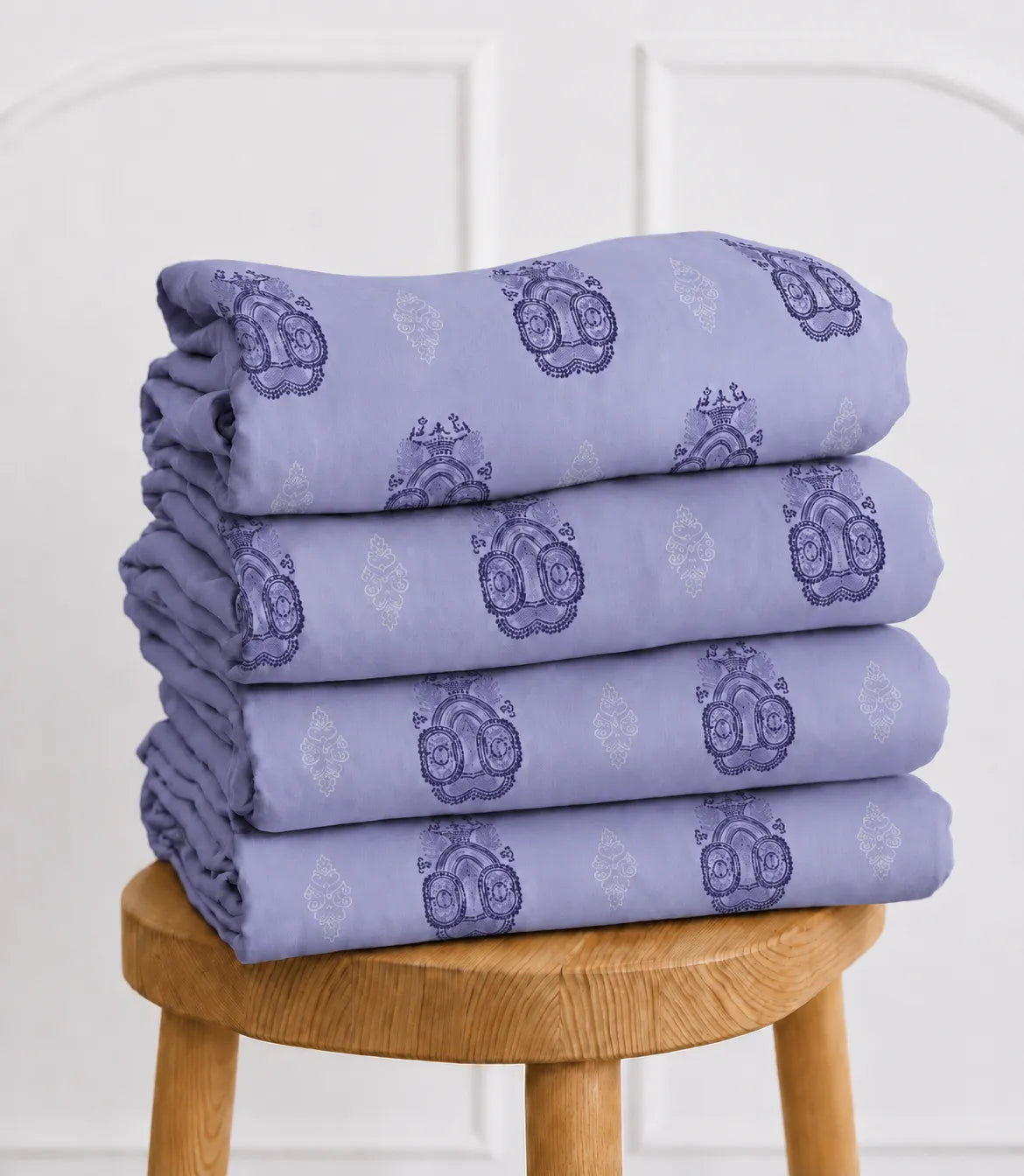 Folded lavender king size cotton bedsheet set with decorative medallion pattern neatly stacked on a wooden stool, showcasing premium cotton fabric and fine craftsmanship.