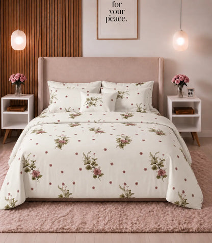Queen-size cotton bedsheet set with a soft ivory base, pink floral bouquets, green leafy accents, and scattered tiny blossoms, neatly styled on a modern upholstered bed in a warm, elegant bedroom.