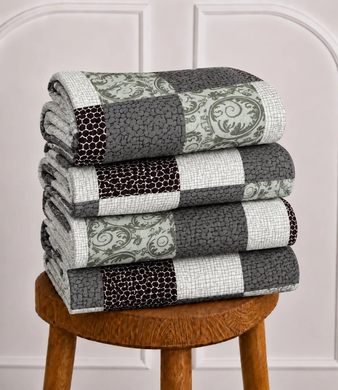 Neatly folded queen size cotton bedsheet showcasing a modern patchwork design with black, grey, sage green, and white geometric squares, stacked on a wooden stool.