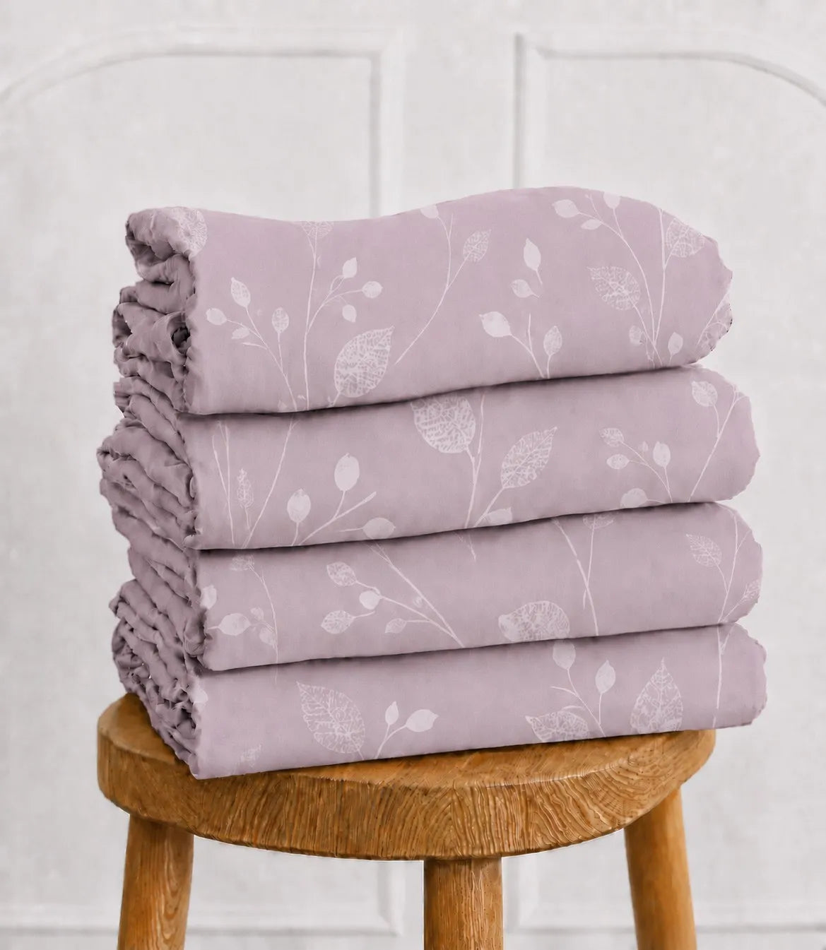 Folded blush lavender cotton bedsheet set with a subtle white leaf and vine pattern, stacked neatly on a rustic wooden stool, highlighting the soft fabric, elegant print, and premium finish.