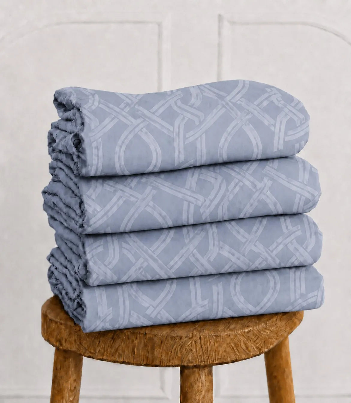 Folded powder blue king size cotton bedsheet with contemporary interwoven geometric print displayed on a wooden stool.