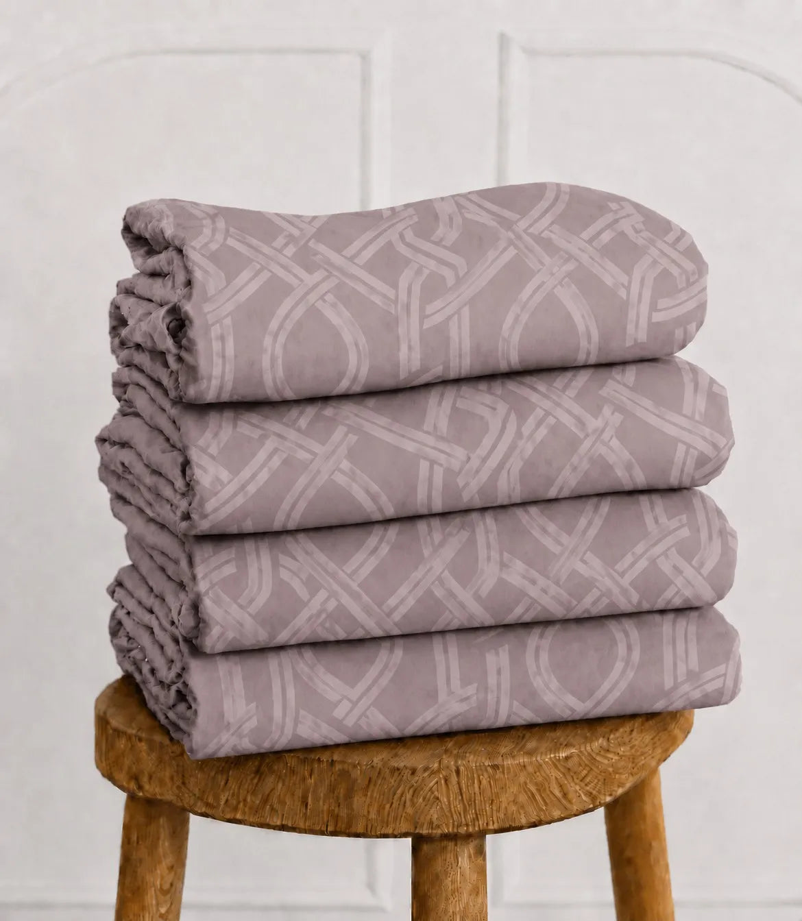 Folded blush mauve cotton bedsheet set with interwoven geometric print stacked on a wooden stool.