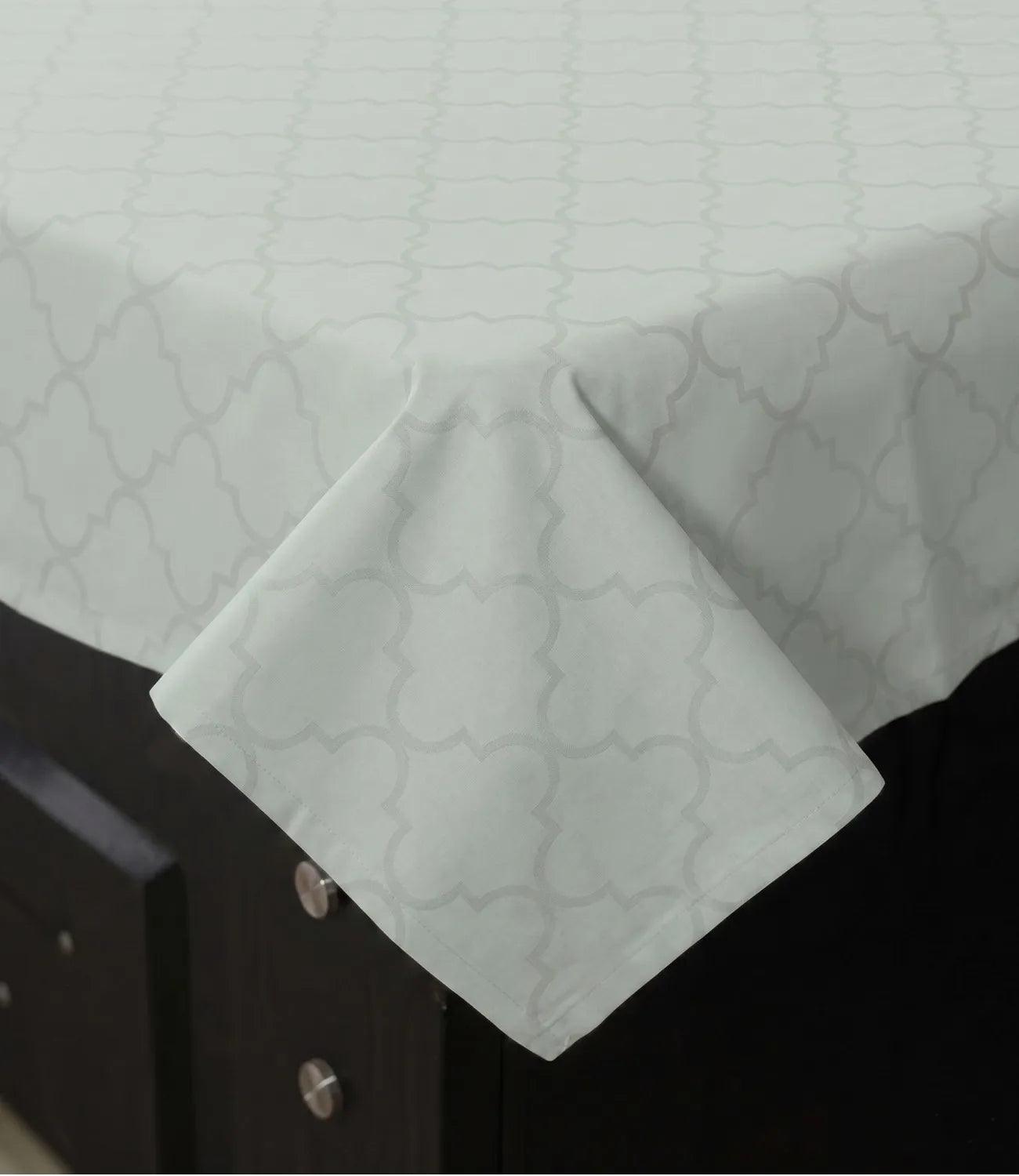 Close-up of a light sage green cotton bedsheet showing the subtle geometric quatrefoil print, fine stitching, and smooth fabric texture.