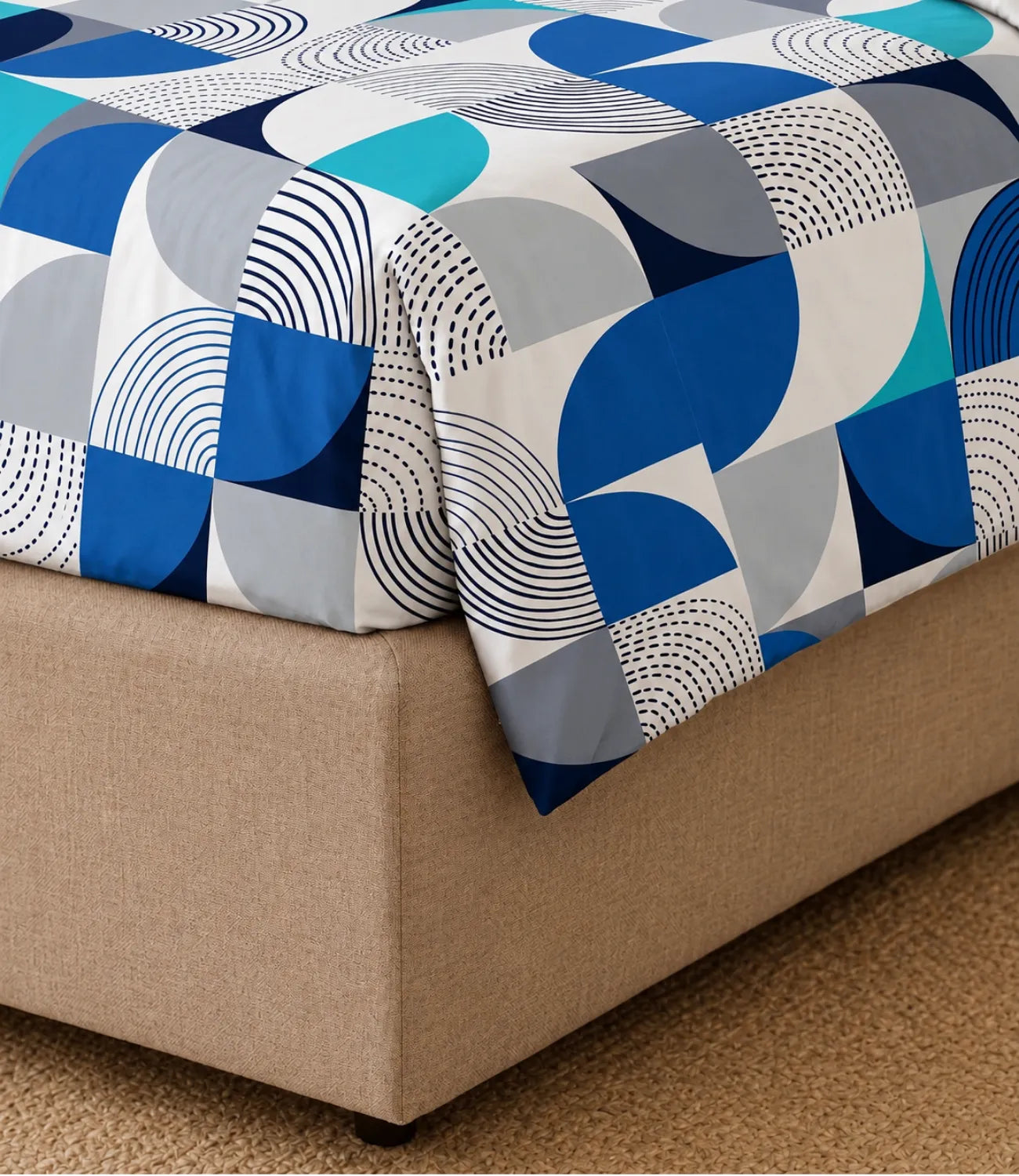 Close-up of a king-size cotton bedsheet with a modern blue, teal, grey, and white geometric pattern draped over the corner of a bed, showcasing fabric texture and print details.